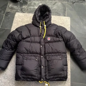 Svart dunjacka från Fjällräven - Säljer en svart dunjacka från Fjällräven i mycket bra skick. Jackan har en mysig huva med gula dragsnören och praktiska fickor framtill. Perfekt för kalla vinterdagar! 🖤