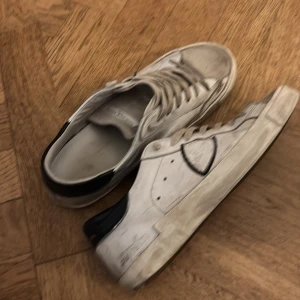 Vita sneakers med svarta detaljer - Säljer ett par vita sneakers från Philippe Model med svarta detaljer. Skorna har snörning och är i acceptabelt skick med synliga tecken på användning. Perfekta för en avslappnad stil och passar till de flesta outfits. Storlek är inte specificerad, storlek 41.