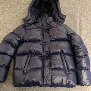 Mörkblå dunjacka från Polo Ralph Lauren - Säljer en snygg mörkblå dunjacka från Polo Ralph Lauren. Den är perfekt för kalla vinterdagar med sin tjocka vaddering och huva. Jackan har en dragkedja framtill och två praktiska fickor. Den är i mycket bra skick och redo för nya äventyr! Storlek 12-14 år liten i storleken så mer 10-12 år. Frakt ca 70 kr.