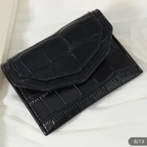 Svart plånbok  - Snygg svart plånbok med krokodilpräglat mönster. Den är kompakt och perfekt för att hålla kort och kontanter. Plånboken har en praktisk knappstängning och är tillverkad i syntetmaterial. 