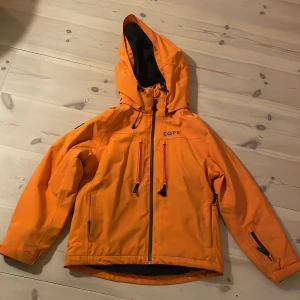 Orange skidjacka från EQPE - Säljer en snygg orange vindjacka från EQPE. Den har en praktisk huva och dragkedja framtill. Ficka på ärmen för liftkort. Snölås i midjan. Lite sliten med färgskiftningar i sömmar runt ärmar och fickor. I övrigt mycket  fin jacka.