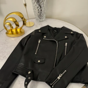 Svart skinnjacka med dragkedjor - Säljer en snygg svart skinnjacka i mycket bra skick. Den har coola dragkedjor och knappar som detaljer, vilket ger den en edgy look. Perfekt för höst och vår, och funkar lika bra till fest som till vardags. Jackan har en klassisk bikerstil med en normal passform.