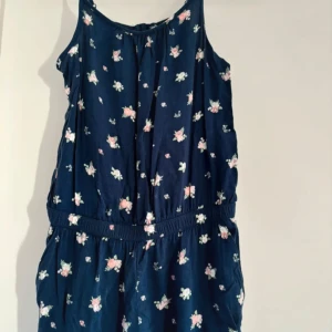 Blommig ärmlös klänning - Söt ärmlös klänning i mörkblått med blommigt mönster. Tillverkad i 100% viskos, vilket gör den lätt och bekväm. Perfekt för sommarens varma dagar eller en avslappnad vårdag. Klänningen har en elastisk midja för en bekväm passform. Är i storlek 164 från HM. 