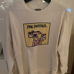 Vit Pink Panther tröja från Zara - Säljer en supercool vit långärmad tröja från Zara med Pink Panther-tryck på framsidan. Perfekt för en avslappnad stil och att visa lite retro-vibbar. Tröjan är i bra skick och passar perfekt till jeans eller shorts. Skön och bekväm för vardagsbruk!