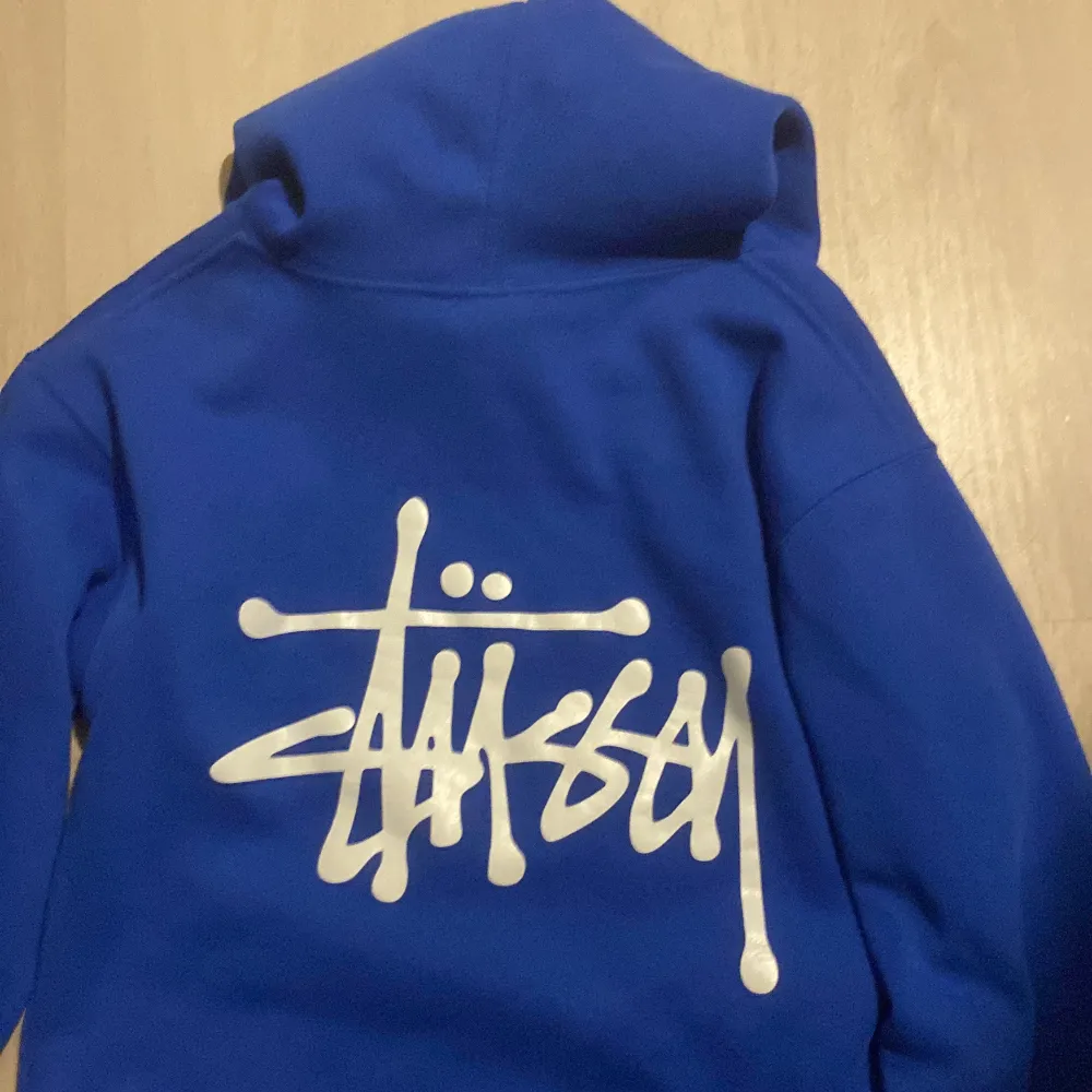 Säljer en snygg blå hoodie från Stussy i mycket bra skick. Den har en stor vit logga på både bröstet och ryggen. Perfekt för en casual look och passar bra till både höst och vår. Den är gjord i mjukt material och har en klassisk känguruficka framtill.. Tröjor & Koftor.