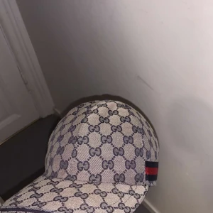 Beige keps med GG-mönster - Säljer en snygg beige keps från Gucci med det klassiska GG-mönstret. Kepsen har en justerbar rem baktill och en röd och blå randdetalj på sidan.  Äkta Gucci keps