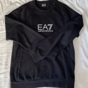 Svart tröja från Emporio Armani EA7 - Säljer en stilren svart tröja från Emporio Armani EA7. Den har en klassisk design med logotypen tryckt på bröstet. Perfekt för både vardag och lite finare tillfällen. Tröjan är i använt skick och passar perfekt till höst och vinter.