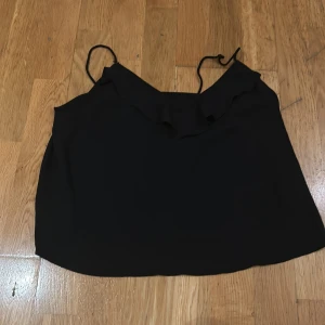 Svart ärmlös topp med volangdetalj från Gina Tricot - Säljer en stilren svart ärmlös topp med tunna axelband och en söt volangdetalj framtill. Perfekt för både vardag och festliga tillfällen. Toppen är i bra skick och passar till många olika outfits! Jag har sytt upp ena snöret och knutit det andra. Ända defekten.