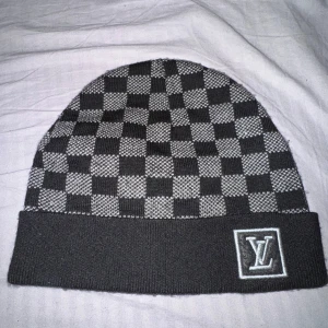 Svart och grå rutig mössa - Snygg svart och grå rutig mössa med ett klassiskt mönster. Perfekt för att hålla värmen under kyliga dagar. Den har en uppvikt kant och en broderad logga framtill. Passar både till vardags och mer uppklädda tillfällen.