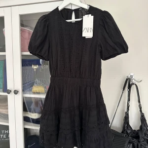Svart klänning från Zara - Säljer en superfin svart klänning från Zara i storlek S. Den har korta puffärmar och ett sött broderat mönster. Perfekt för både vardag och fest! Klänningen är i nytt skick och har en bekväm passform med en lätt volangdetalj i nederkant. Helt oanvänd med lappen kvar!