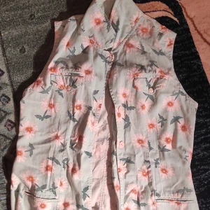 Blommig väst med knappar - Säljer en söt blommig väst med knappar framtill. Västen har ett härligt mönster med rosa blommor och gröna blad på en ljus bakgrund. Perfekt för vår och sommar! 🌸