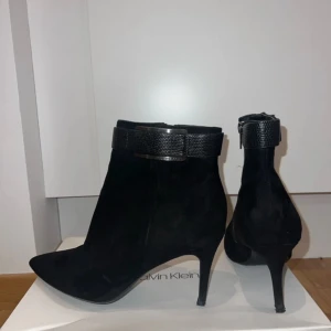 Svarta stövletter från Calvin Klein Strl 40. - Snygga svarta stövletter i mocka från Calvin Klein med spetsig tå och klack. De har en elegant dragkedja på sidan och en dekorativ rem runt ankeln. Perfekta för höst och vinter, både till vardags och fest. Använda men i mycket bra skick. Storlek 40.