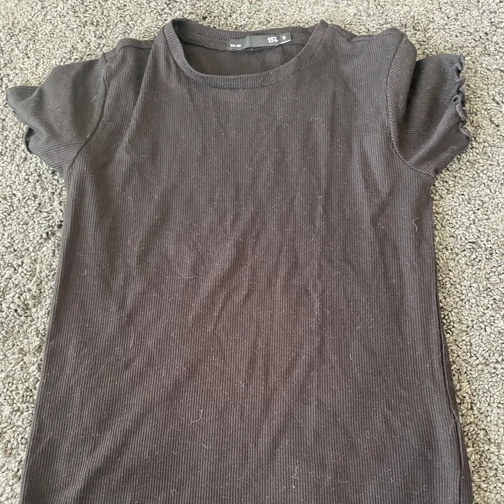 Säljer en svart ribbad topp i storlek S. Den har korta ärmar med volangdetaljer och en enkel rund halsringning. Perfekt för vardagsbruk och kan enkelt matchas med jeans eller kjol.. Yläosat.