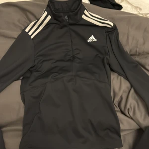Svart långärmad tröja från Adidas - Säljer en svart långärmad tröja från Adidas i mycket bra skick. Den har vita ränder på axlarna och Adidas-loggan på bröstet. Perfekt för träning eller vardagsbruk. Materialet är Primegreen, vilket är hållbart och bekvämt. Passar bra till höst och vår.