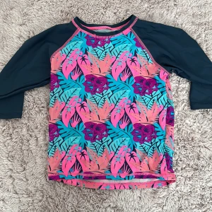 Färgglad rashguard med tropiskt mönster - Säljer en supercool rashguard med tropiskt mönster i rosa, blått och lila. Perfekt för stranden eller poolen! Den har trekvartsärmar och är gjord i ett snabbtorkande material. Passar perfekt för sommarens alla äventyr! 🏝️🐚, den har inget varu mäske eller storlek men jag kan gissa att den är 8-9