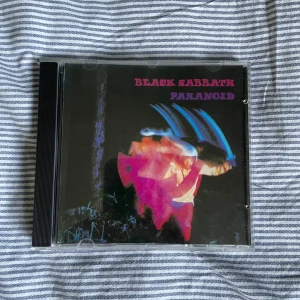 Black Sabbath - Paranoid CD - Säljer en klassisk CD av Black Sabbath med albumet 'Paranoid'. Omslaget har en mörk design med en färgglad figur i rörelse. Skivan innehåller låtar som 'War Pigs', 'Paranoid' och 'Iron Man'. Perfekt för rockälskare och samlare!