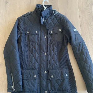 Svart quiltad jacka från Barbour - Säljer en snygg svart quiltad jacka från Barbour International. Den har en stilren design med knappar och dragkedja framtill, samt en hög krage med spänne. Jackan har fyra fickor med knappar och är perfekt för höst och vår. Den är i mycket bra skick och redo för nya äventyr!
