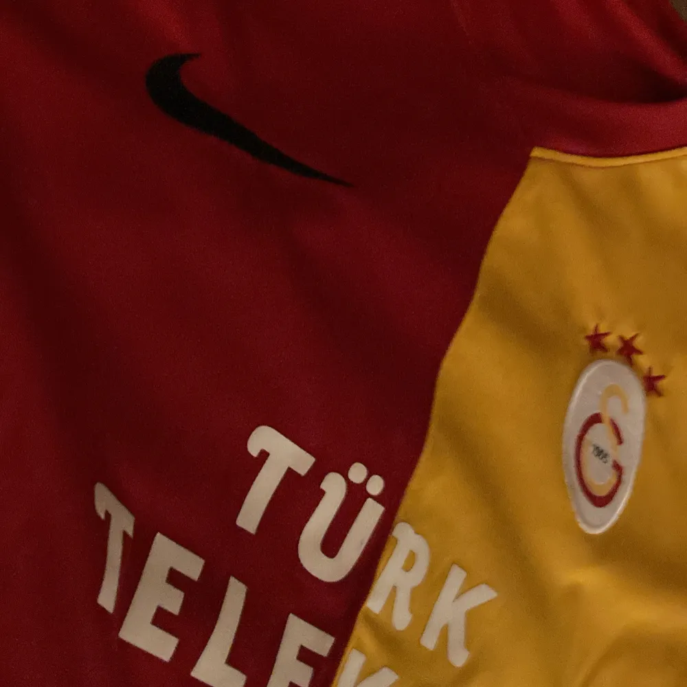 Säljer en Galatasaray fotbollströja från Nike i rött och gult. Den är kortärmad och har sponsorloggor från Türk Telekom och Avea. Tröjan är i mycket bra skick och har en autentisk etikett. Perfekt för fans som vill visa sitt stöd! Passar för 10-12 åringar.. T-shirts.