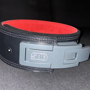 Svart och rött lyftarbälte från SBD - Säljer ett robust lyftarbälte från SBD i svart och rött. Bältet har en kraftig metallspänne och är perfekt för styrketräning. Insidan är röd och utsidan svart med snygga sömmar. Perfekt för att ge extra stöd vid tunga lyft! Helt ny storlek M Priset är inte hackat i sten