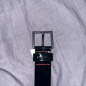 Svart bälte med röd söm - Säljer ett svart bälte med en snygg röd söm. Bältet har en klassisk design med ett fyrkantigt spänne. Perfekt för att ge en stilren touch till din outfit. Det finns några repor på bältet, men det är fortfarande i bra skick. Passar både till vardags och fest!