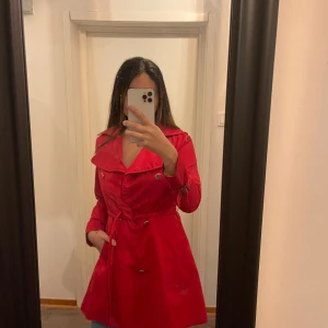 Röd jacka/kappa från H&M - Säljer en snygg röd trenchcoat från H&M i storlek XS. Jackan har långa ärmar och ett bälte i midjan för en stilren look. Perfekt för höst och vår! 🧥