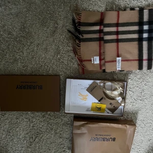 Burberry halsduk - Säljer en klassisk beige rutig halsduk från Burberry. Den är i nytt skick och kommer med originalförpackning. Perfekt för att hålla sig varm och stilren under kyliga dagar. Den har fransar i ändarna och det ikoniska Burberry-mönstret i svart och rött.
