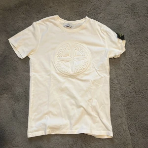 Vit t-shirt från Stone Island - Säljer en snygg vit t-shirt från Stone Island i mycket bra skick. Den har ett stort, broderat Stone Island-märke på framsidan som ger en cool look. Perfekt för både vardag och fest! Passar bra till jeans eller shorts. Inga fläckar eller hål.