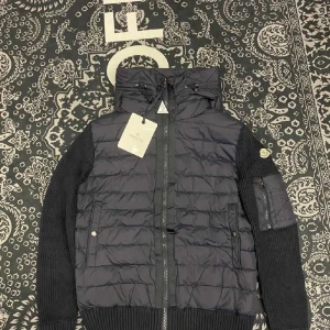 Svart jacka från Moncler - Säljer en snygg svart jacka från Moncler i mycket bra skick. Den har en quiltad design med stickade ärmar och en praktisk huva. Perfekt för höst och vinter! Jackan har dragkedja och en liten logga på ärmen. Passar både till vardags och lite finare tillfällen. Den har även nfc Scan 