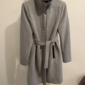 Grå kappa med bälte - Säljer en stilren grå kappa i mycket bra skick. Den har en elegant design med ett bälte i midjan och dragkedjor vid fickorna. Perfekt för höst och vår! Passar både till vardags och festliga tillfällen.