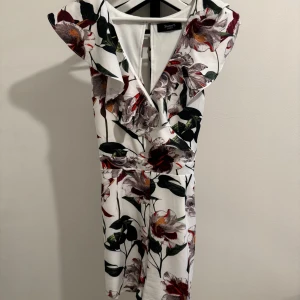 Blommig klänning från Sisters Point - Säljer en superfin blommig klänning från Sisters Point. Den är vit med stora blommor i rött, grönt och lila. Klänningen har en v-ringad design och volangdetaljer. Den är ärmlös och har ett knytband i midjan för en snygg passform. Perfekt för sommarens fester eller en dag på stan!