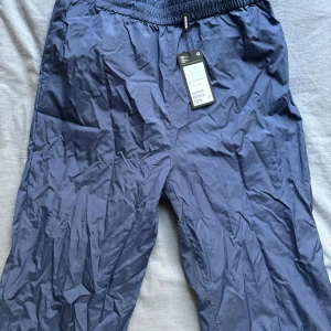 Blå Windbreaker Byxor från Weekday - Ett par blåa windbreakers/parachute byxor från weekday Aldrig använda, med prislapp kvar, inga defekter Fick som present några år sedan, säljer då de nu är för små (L: 100cm, W: 38cm, Innersöm: 70cm, Benöppning: 14cm)