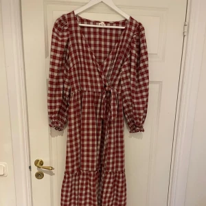 Rutig röd och vit klänning - Charmig långklänning i röd och vit rutig design. Klänningen har en omlottstil med knytband i midjan och långa ärmar med elastiska muddar. Perfekt för höst och vår med sitt mjuka och bekväma material.