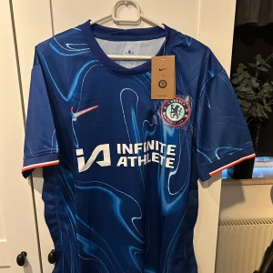 Blå Chelsea fotbollströja - Säljer en ny Chelsea fotbollströja i blått med ett coolt mönster. Tröjan är kortärmad och har Nike-loggan samt 'Infinite Athlete' tryckt på framsidan. Perfekt för alla Chelsea-fans! 💙