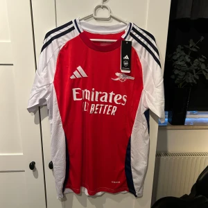 Röd och vit fotbollströja från Adidas - Säljer en ny Adidas fotbollströja i rött och vitt med korta ärmar. Den har snygga mörkblå detaljer och ett tryck av 'Emirates Fly Better' på framsidan. Perfekt för alla Arsenal-fans! Tröjan är i nytt skick med etikett kvar.