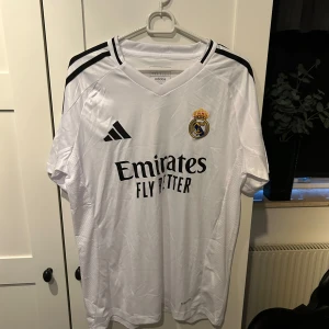 Vit Real Madrid fotbollströja från Adidas - Säljer en vit Real Madrid fotbollströja från Adidas i nytt skick. Tröjan har korta ärmar och klassiska svarta ränder på axlarna. Den är gjord i ett lätt och bekvämt material, perfekt för både träning och match. Real Madrid-loggan och sponsortrycket 'Emirates Fly Better' pryder framsidan. Perfekt för alla fans! ⚽