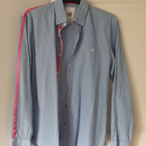 Blå och vit randig skjorta från Mouli - Säljer en snygg blå och vit randig skjorta från Mouli. Skjortan har långa ärmar och en cool rosa detalj längs med ärmen. Den har knappar framtill och en liten broderad logga på bröstet. Perfekt för kalas och lite finare tillfällen! Använd 2-3 gånger med ett nypris på 899 kr!!