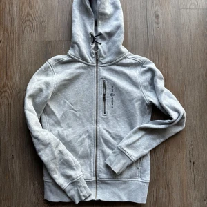 Grå hoodie från Sail Racing - Säljer en snygg och bekväm grå hoodie från Sail Racing i storlek S. Den har en dragkedja framtill och en justerbar huva med snörning. Perfekt för en avslappnad stil eller som ett extra lager under kyliga dagar. Den är i bra skick och redo för nya äventyr!