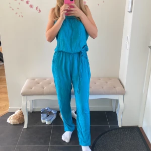 Turkos byxdress - Säljer en snygg turkos byxdress som är perfekt för sommaren. Den har en avslappnad passform med knytband i midjan för att justera passformen. Materialet ser lätt och luftigt ut, vilket gör den idealisk för varma dagar. Perfekt för både vardag och fest! Färgen är mer i petrol i verkligheten