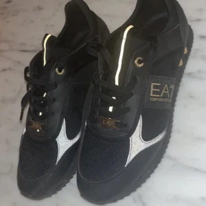 Svarta sneakers från Emporio Armani - Snygga svarta sneakers från Emporio Armani med silverdetaljer och snörning. Skorna har en sportig design med märkets logga på sidan och en bekväm sula.