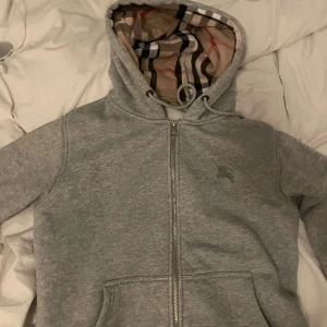 Grå hoodie från Burberry - Säljer en stilren grå hoodie från Burberry med dragkedja och klassiskt rutigt mönster i huvan. Tröjan har en broderad logga på bröstet och är perfekt för en avslappnad look. Passar bra till både jeans och joggers.