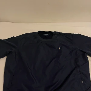 Mörkblå Vindjacka från Polo Golf Ralph Lauren - Säljer en stilren mörkblå vindjacka från Polo Golf Ralph Lauren. Tröjan är tillverkad i 100% polyester och har en liten broderad logotyp på bröstet. Perfekt för en avslappnad och sportig look. fickor och dragkedja nästan vid sidan av jackan.