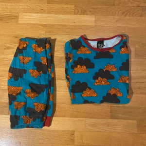 Mönstrad pyjamas från Villervalla - Säljer en färgglad pyjamas från Villervalla med ett unikt mönster av svarta och orangea moln på en blå bakgrund. Pyjamasen har långa ärmar och byxben med röda muddar vid ärmslut och benslut. Perfekt för mysiga kvällar! Väldigt bra skick och perfect för barn. Namnlös. 100kr. Priset är diskuterbart! 