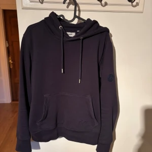 Mörkblå hoodie från Strömberg - Säljer en stilren mörkblå hoodie från Strömberg i bomull. Den har en klassisk design med känguruficka och justerbar huva med snören. Perfekt för en avslappnad look.
