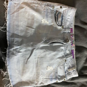 Ljusblå lågmidjad jeanskjol med slitningar - Trendig ljusblå jeanskjol från Miss Sixty med slitna detaljer och fransig kant. Kjolen har klassisk femficksdesign och dekorativa sömmar på bakfickorna. Perfekt för en avslappnad stil. Denna kjol är även lågmidjad, passar både M och S