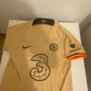 Chelsea FC beige  - Säljer en beige Chelsea FC fotbollströja från Nike med korta ärmar. Tröjan har klubbens logga och sponsortryck på framsidan samt spelarnamnet Kanté och nummer 7 på baksidan. Tillverkad i Dri-FIT-material för optimal komfort.