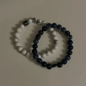 Två elastiska armband med pärlor - Säljer två elastiska armband med pärlor. Ett armband har vita pärlor med grå marmorering och ett metalliskt inslag, medan det andra har mörkblå pärlor med ett liknande metalliskt inslag. Perfekt till en snygg outfit speciellt till sommaren. Det blåa armbandet är lite sönder, två av pärlorna har gått av på mitten då de redan var sprickor när jag köpte armbandet, finns inga sprickor kvar. Gå även att bara köpa det vita. Kan gå att förhandla
