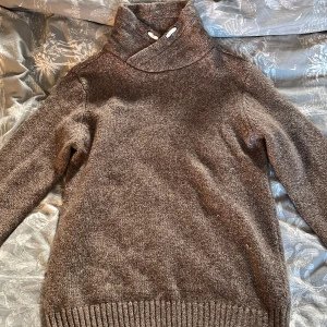 Brun stickad tröja med hög krage - Mysig brun stickad tröja med hög krage och dekorativa knappar. Perfekt för kyliga dagar med sin varma och bekväma design. Ribbstickade muddar vid ärmslut och nederkant.