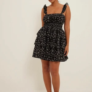 Svart blommig klänning från NA-KD - Charmig svart klänning från NA-KD med små vita blommor. Den har volanger och en smockad midja för en smickrande passform. Klänningen är ärmlös med breda axelband och en söt knytning framtill. Perfekt för en somrig look! 🌸