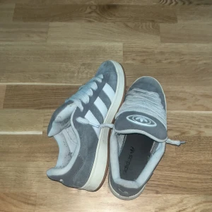 Adidas campus - Säljer ett par klassiska grå och vita Adidas sneakers med de ikoniska tre ränderna på sidan. Skorna har snörning och en bekväm sula, perfekt för vardagsbruk. 