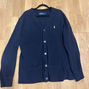 Mörkblå kofta från Ralph Lauren - Säljer en stilren mörkblå kofta från Ralph Lauren med knappar framtill och två fickor. Den har en broderad logga på bröstet och långa ärmar. Perfekt för en klassisk look.